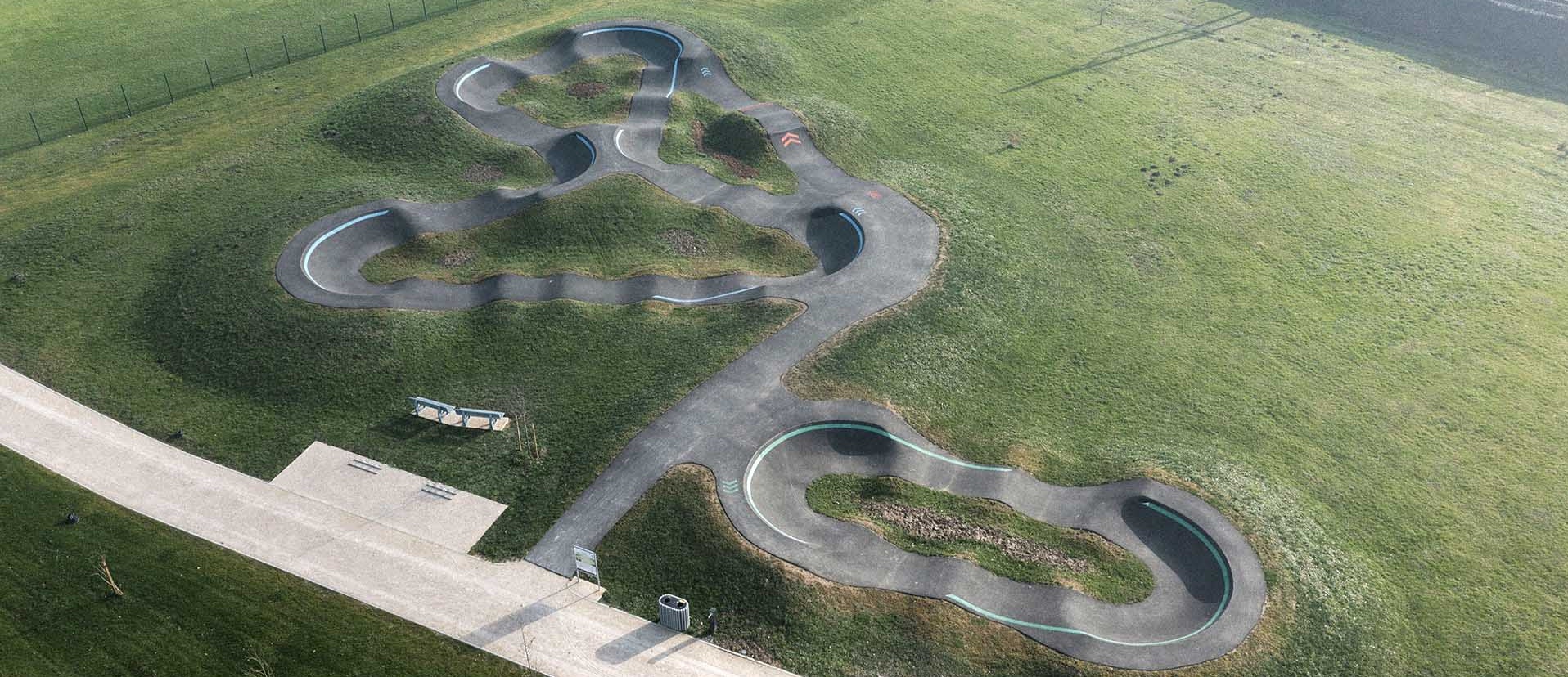 Vue aérienne d'un pumptrack moderne en enrobé dessiné sur une pelouse verte à côté d'une voie ferrée.
