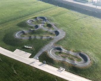 Aerial view of a modern asphalt pumptrack with looping tracks on a green lawn next to a railway line.