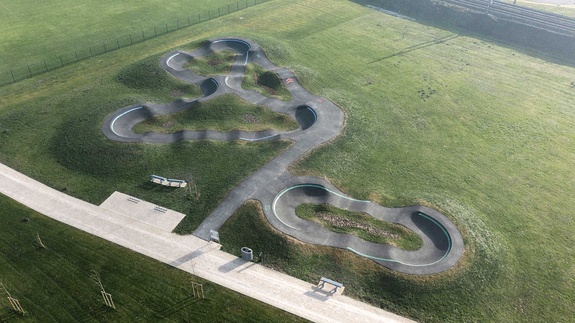 Aerial view of a modern asphalt pumptrack with looping tracks on a green lawn next to a railway line.