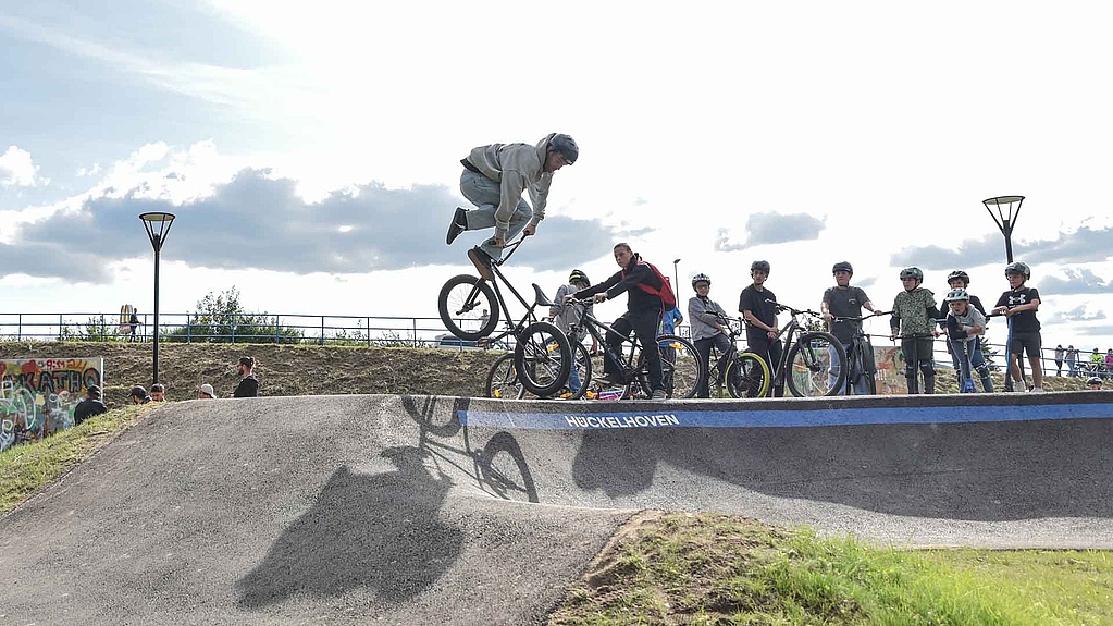 BMX-Fahrer springt über den Pumptrack im Urban Sports Park Hückelhoven, Zuschauer beobachten das Event.