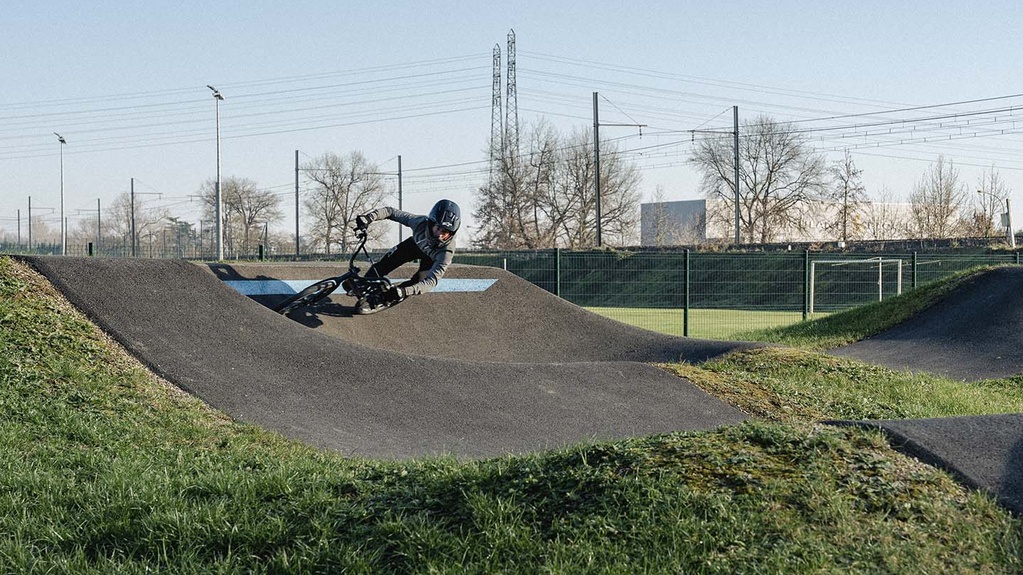 A cyclist in protective gear riding through a banked turn on an outdoor asphalt pumptrack track.