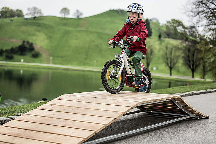 Kinder mit roter Jacke fährt über Bike Obstacle mit See und Grashügel im Hintergrund