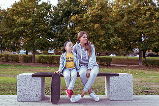 Mutter sitzt mit Kind und Skateboard auf einer Parkbank