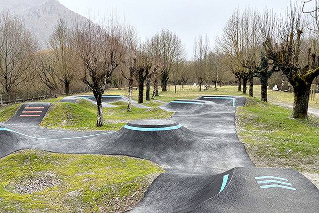 Bodenperspektive der Asphalt-Wellen und Kurven des Pumptracks in Saint-Béat-Lez mit roten Richtungspfeilen.