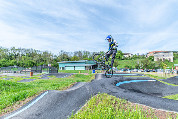 Un jeune rider en BMX effectue un saut aérien au-dessus d'une bosse sur un pumptrack en extérieur.