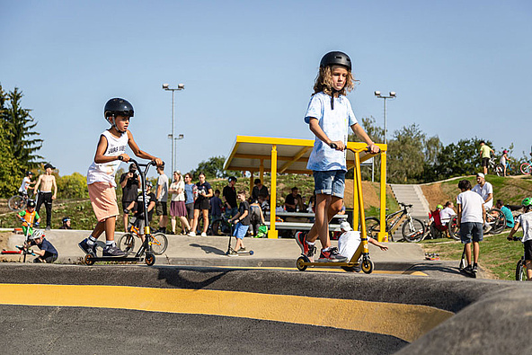 [Translate to Französisch:] Scooterfahrer am Rand eines Pumptrack mit vielen Menschen im Hintergrund