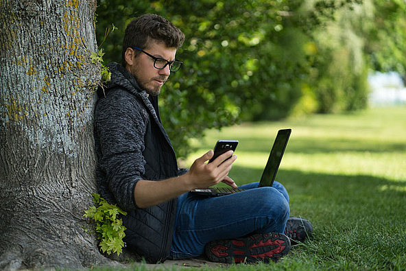 [Translate to Französisch:] You sit in the park by the tree with laptop and smartphone
