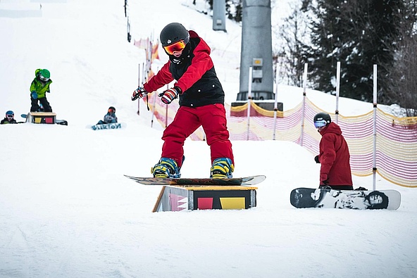 Kind mit Snowboard fährt über eine Box und weitere Snowboarder im Hintergrund