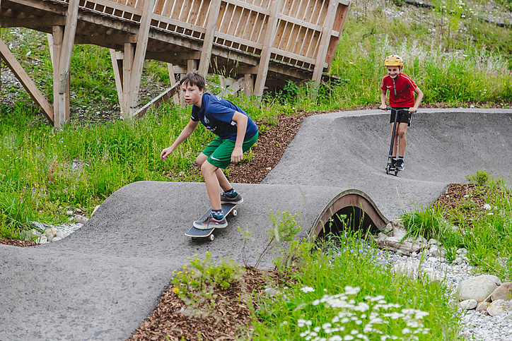 Zwei Kinder fahren mit Skateboard und Scooter im Pumptrack